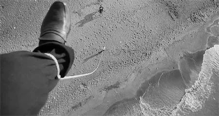 Black-and-white film still of a shoe suspended above a beach and breaking waves.